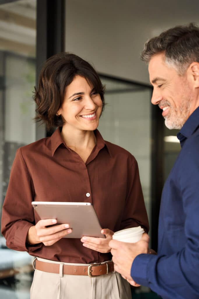 Happy professional business man and woman colleagues having conversation at work. Executive team of two business people working talking using tablet tech in corporate office. Vertical candid photo.